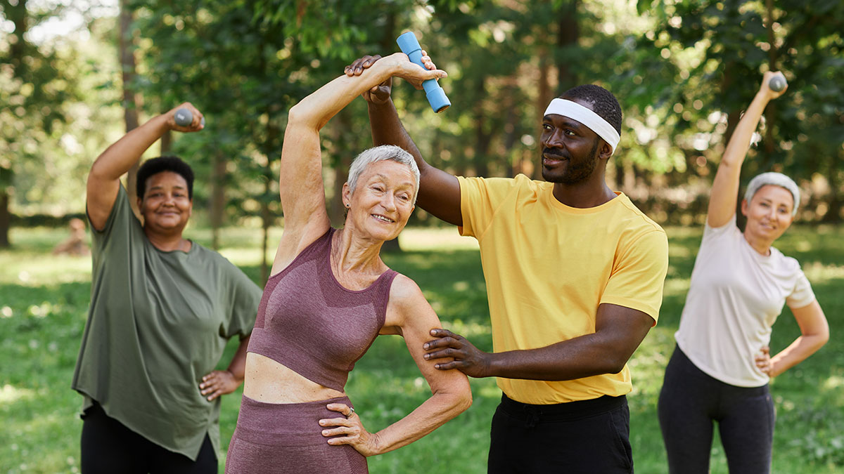 A group of senior citizens exercising in a grassy area, assisted by a trainer.