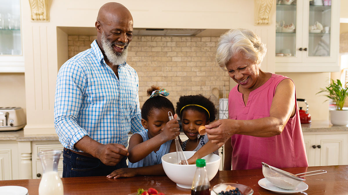 An elderly Black couple in their kitchen mixing ingredients in a bowl with their grand daughters.