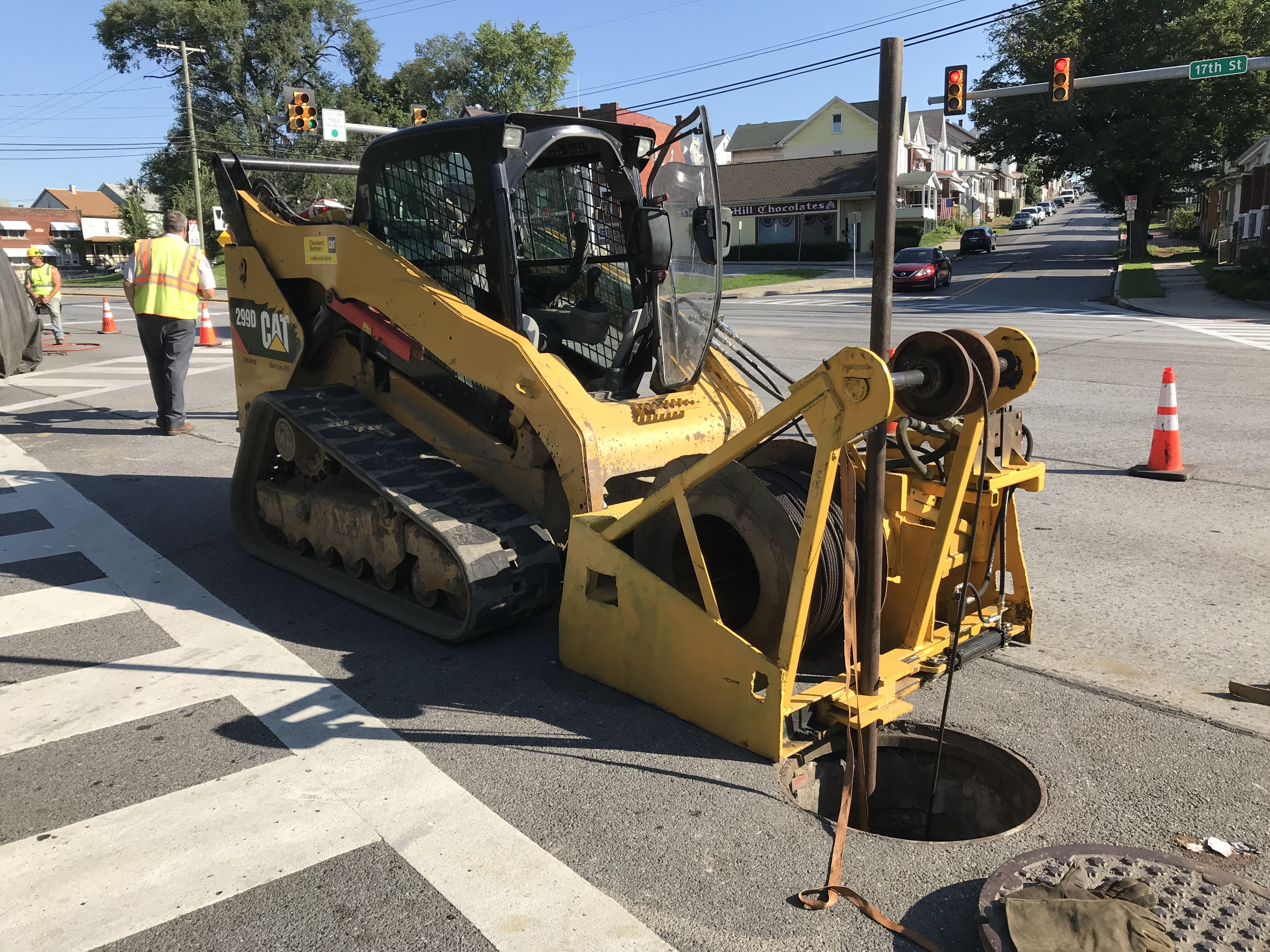 An image of a yellow piece of equipment with a pulley system on the front setting up at an open manhole cover preparing to pull the white PVC liner through the pipe