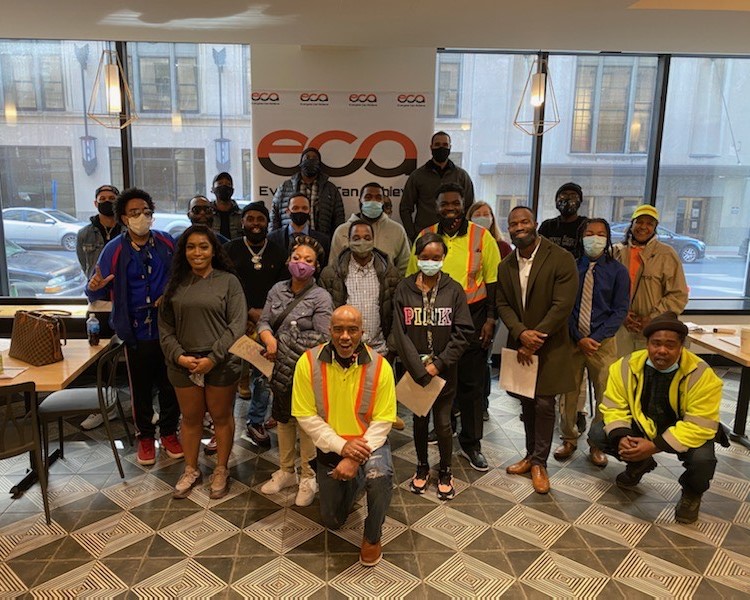 An image of a group of individuals standing in rows in front of an Everyone Can Achieve (ECA) Corp banner with large windows and a city street with cars in the background