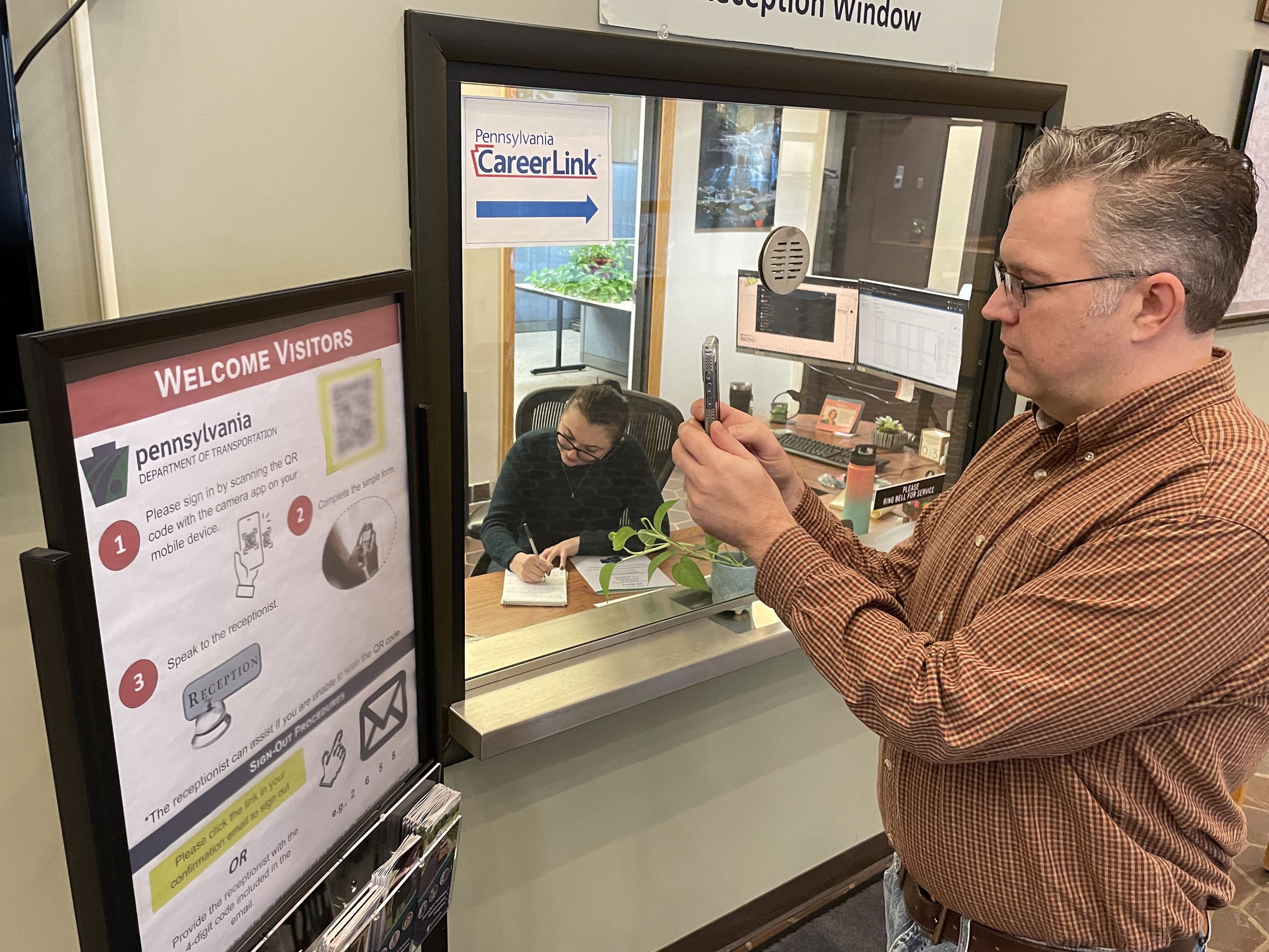 An individual wearing a brown plaid shirt and blue jeans uses a mobile device to scan a QR code on a poster hanging outside a receptionist's window while a receptionist sitting at a desk behind the window writes with a pen in a notebook